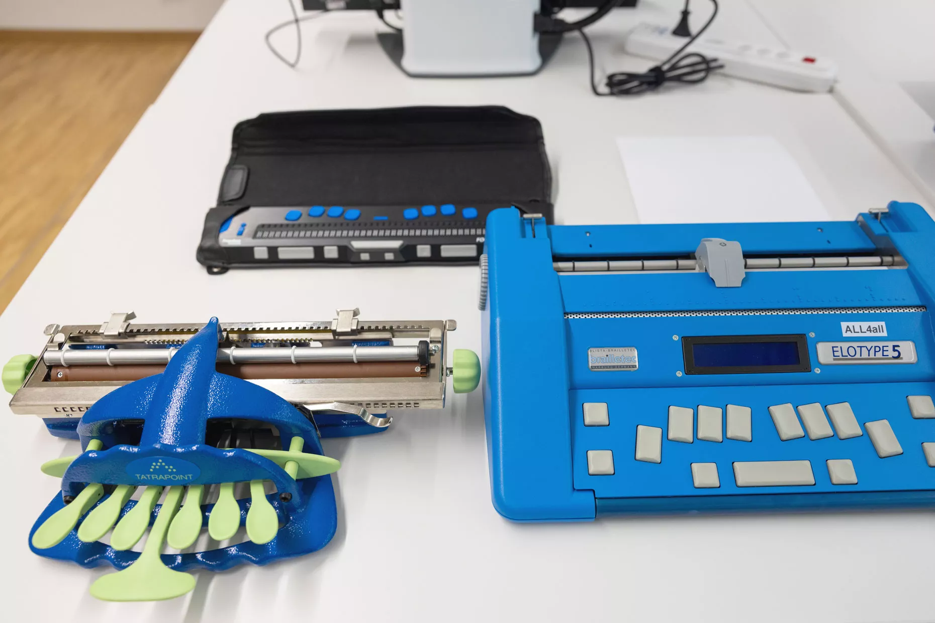 Auf einem Tisch sind drei Geräte aufgebaut. Links eine alte mechanische Braille-Schreibmaschine, rechts eine moderne elektronische Braille-Schreibmaschine und in der Mitte eine Braille-Zeile.